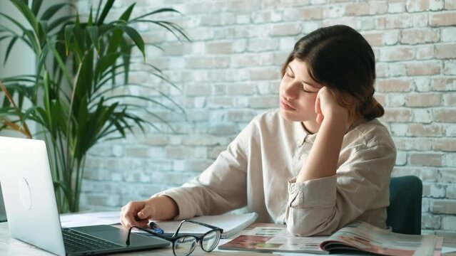 Young woman student sitting at desk full with books textbooks looking at computer screen to unfinished homework feels tired bored and unmotivated, girl loaded with tasks, work backlog overflow concept