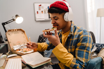 Middle-eastern teenage boy eating pizza studying at home by computer