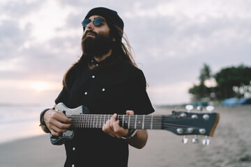 Solo singer in stylish clothes performing rock'n'roll music during hobby recreation at seashore coastline, talented composer creating sound melody on acoustic six stringed guitar - ukulele equipment