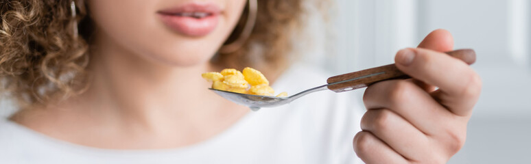 partial view of curly woman holding spoon with tasty corn flakes, banner.
