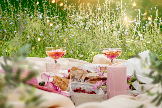 Aesthetic Summer Sunny Lunch Picnic With Baguette, Flowers, Cocktail And Berries At Pale Beige Blanket At Grass Outdoor Background With Sunbeam. Front View.