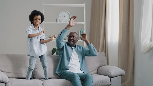 Funny adult loving father sits in room plays with daughter kid girl cheerful jumping on couch dancing rhythmic music dad supports child having fun at home on weekend dance together makes comical move