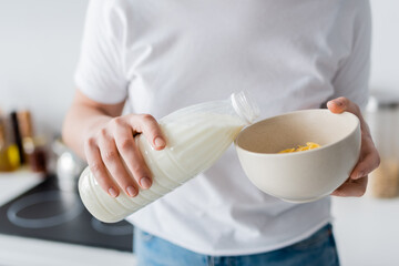 cropped view of blurred woman pouring fresh milk into bowl with corn flakes.
