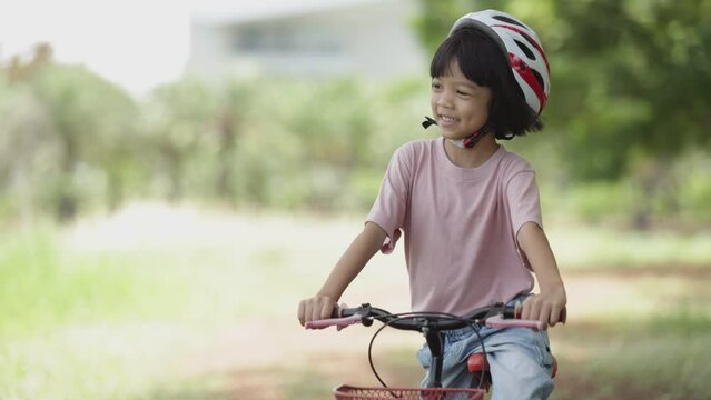 Thai Asian kid girl, aged 4 to 6 years old, looks cute, wears a helmet. and a bicycle She is play riding a bicycle to exercise in the park. It is a fun outdoor activity and learning about nature.