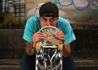 Portrait of a young latin skater sitting and holding his board while looking at the camera. Urban lifestyle concept. © Jorge Sion