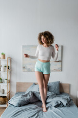full length of happy young woman in white pajamas standing on bed at home.