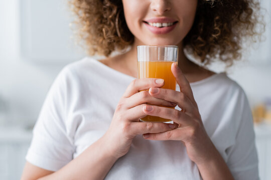 cropped view of curly smiling woman holding glass of fresh orange juice.