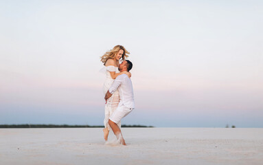 Fototapeta na wymiar A bearded man takes on his hands a smiling blond woman on a background of white sand and bright sky. Portrait of emotional people at sunset. Love in the desert newlyweds. The love story of lovers.