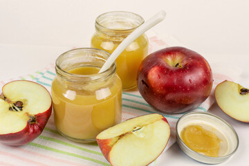 Baby food. Two glass jars with baby apple puree with a spoon and red ripe apples on a light cloth napkin. The first fruit lure of the baby.