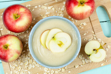 Oatmeal porridge for the baby in a blue bowl, red juicy apples on a wooden board. Baby's first complementary food, baby nutrition.