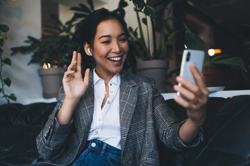 Happy Asian female in electronic earbuds waving during video communication via cellphone...