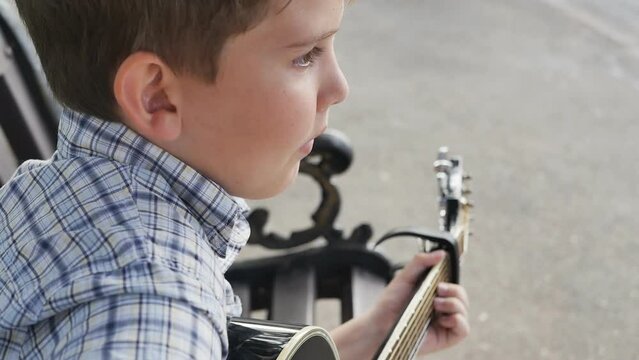 cute european teenager playing acoustic guitar sitting on a street bench close to the road and pavement. The boy earns money by playing a melody. slow motion