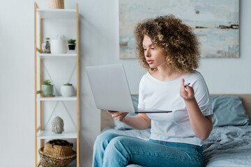 Fototapeta na wymiar curly woman in t-shirt and jeans sitting with laptop on bed at home.