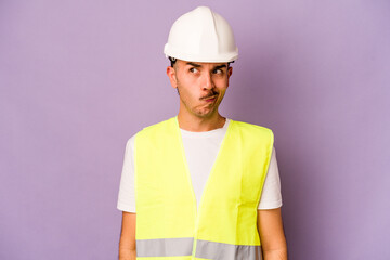 Young hispanic worker man isolated on purple background confused, feels doubtful and unsure.