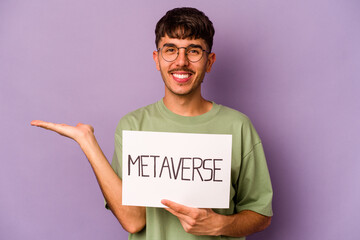 Young hispanic man holding metaverse placard isolated on purple background showing a copy space on a palm and holding another hand on waist.