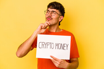 Young hispanic man holding crypto money placard isolated on yellow background looking sideways with...