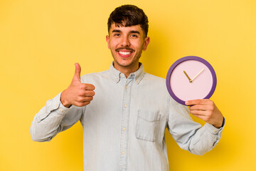Young hispanic man holding a clock isolated on yellow background smiling and raising thumb up