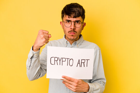 Young Hispanic Man Holding Crypto Art Placard Isolated On Yellow Background Showing Fist To Camera, Aggressive Facial Expression.