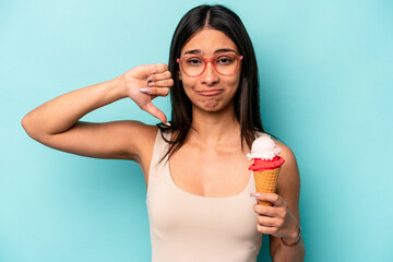 Young hispanic woman holding an ice cream isolated on blue background showing a dislike gesture,...