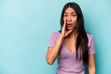 Young hispanic woman isolated on blue background is saying a secret hot braking news and looking...