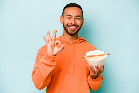 Young hispanic woman eating noodles isolated on blue background cheerful and confident showing ok gesture.