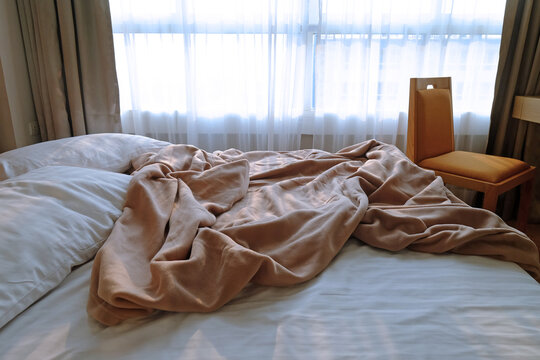 Unmade bed in a bedroom, king size, closed to the window behind the curtain and a comfortable chair 