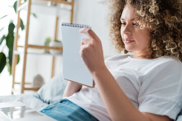 positive woman with curly hair looking at notebook on bed at home.