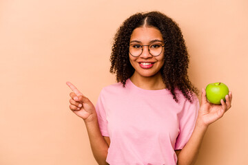 Young African American woman holding an apple isolated on beige background smiling and pointing aside, showing something at blank space.