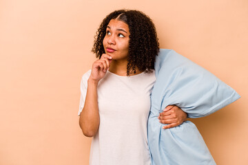 Young African American woman holding a pillow isolated on beige background looking sideways with...