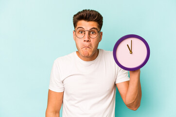 Young caucasian man holding a clock isolated on blue background shrugs shoulders and open eyes...