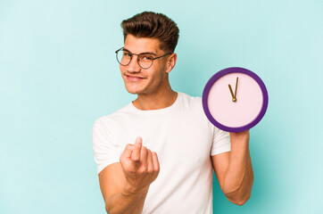Young caucasian man holding a clock isolated on blue background pointing with finger at you as if...