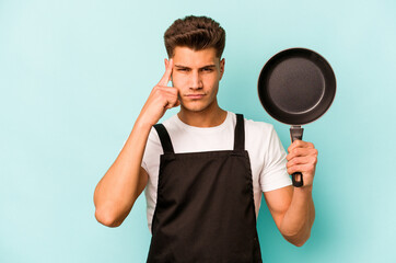 Young caucasian cooker holding frying pan isolated on blue background pointing temple with finger, thinking, focused on a task.