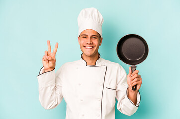 Young caucasian chef man holding flying pan isolated on blue background showing number two with fingers.