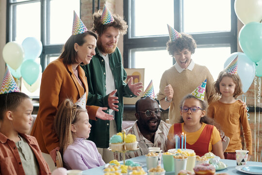 Little Girl In Party Hat Blowing Candles On Birthday Cake With Her Parent And Friends Congratulating Her With Birthday