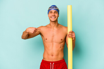 Young caucasian swimmer man holding foam stick isolated on blue background person pointing by hand...