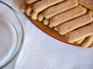 Cropped view of ladyfinger savoiardi biscuit cookies on a wooden cutting board on white tablecloth background with blurred steel bowl and glass plate. Close up