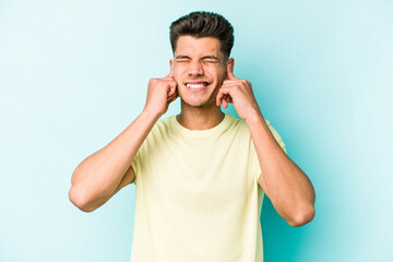 Young caucasian man isolated on blue background covering ears with fingers, stressed and desperate by a loudly ambient.