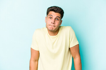 Young caucasian man isolated on blue background shrugs shoulders and open eyes confused.