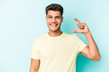 Young caucasian man isolated on blue background holding something little with forefingers, smiling and confident.