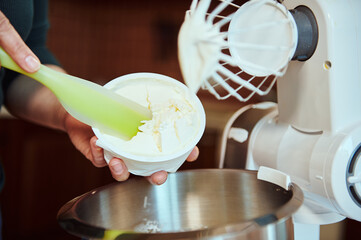 Details. Pastry chef's hands using a silicone spatula to put mascarpone cheese into a steel bowl against a blurred food processor whisk. Preparing traditional Italian Tiramisu