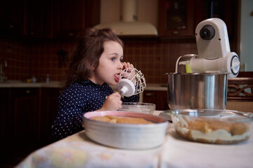 Adorable European little girl licks whisk with leftover sweet whipped cream while her mom is preparing tiramisu dessert at home kitchen
