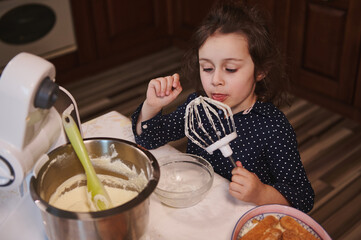 Adorable Caucasian little girl licking sweet delicious whipped cream from the food processor mixer...