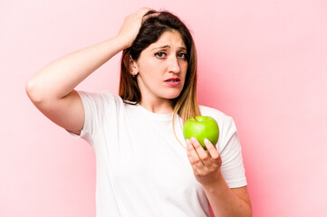 Young caucasian woman holding an apple isolated on pink background being shocked, she has remembered important meeting.