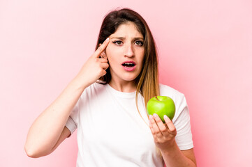 Young caucasian woman holding an apple isolated on pink background showing a disappointment gesture...