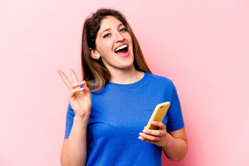 Young caucasian woman holding mobile phone isolated on pink background joyful and carefree showing a peace symbol with fingers.