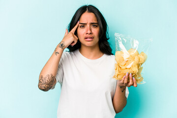 Young hispanic woman holding a bag of chips isolated on blue background showing a disappointment gesture with forefinger.