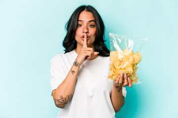 Young hispanic woman holding a bag of chips isolated on blue background keeping a secret or asking for silence.