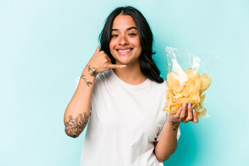 Young hispanic woman holding a bag of chips isolated on blue background showing a mobile phone call...