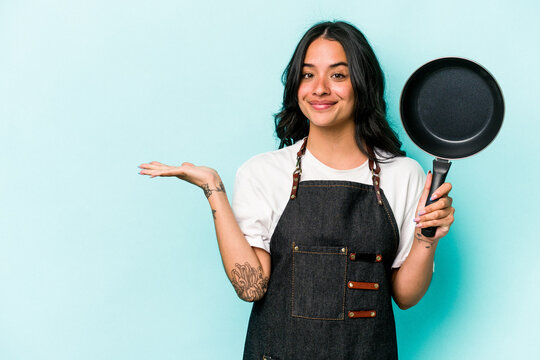 Young hispanic cooker woman holding frying pan isolated on blue background showing a copy space on a palm and holding another hand on waist.