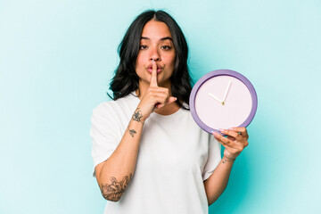 Young hispanic woman holding a clock isolated on blue background keeping a secret or asking for silence.